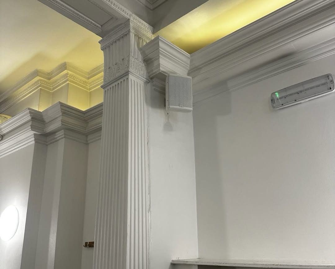 White ornate columns and moulding in a well-lit room at the Mayfair Islamic Centre, with a wall-mounted speaker and an emergency light visible on the right side. Yellow lighting highlights the ceiling details, making this space an inspiring case study in elegant design for an Islamic Centre. White ornate columns and moulding in a well-lit room at the Mayfair Islamic Centre, with a wall-mounted speaker and an emergency light visible on the right side. Yellow lighting highlights the ceiling details, making this space an inspiring case study in elegant design for an Islamic Centre.