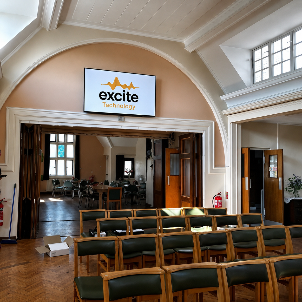 A room with rows of wooden chairs faces a screen displaying the excite Technology logo at Merton Park Baptist Church. Doors at the back open to another room with more chairs and tables, while sunlight streams through the windows.