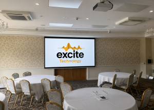 Conference room with round tables and chairs, a large screen displaying the excite Technology logo, and neutral decor with patterned wallpaper and carpet, located within the elegant Shendish Manor Hotel & Country Club—perfect for combining business meetings with a round of golf.
