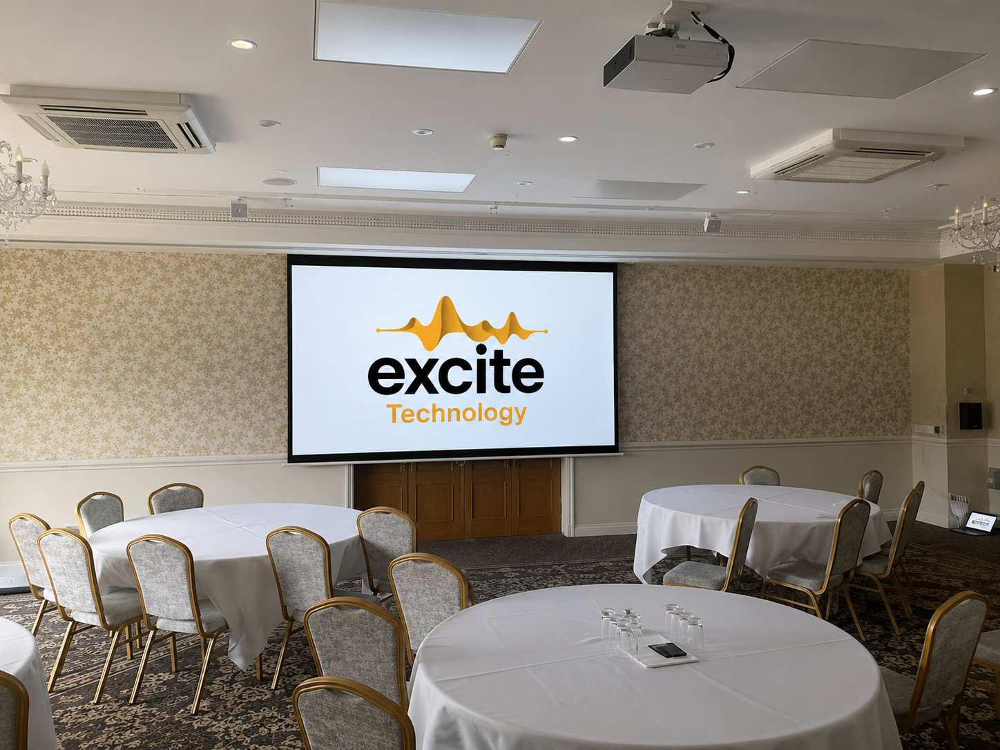 Conference room with round tables and chairs, a large screen displaying the excite Technology logo, and neutral decor with patterned wallpaper and carpet, located within the elegant Shendish Manor Hotel & Country Club—perfect for combining business meetings with a round of golf.