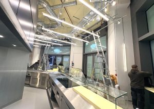 Several people set up a modern shop interior for ATIS Foods in London, with ladders, glass display cases, and exposed pipes on the ceiling. Bright lights illuminate the space.