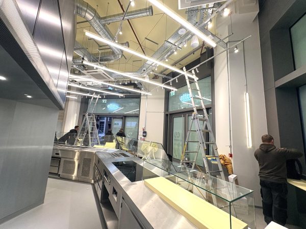 Several people set up a modern shop interior for ATIS Foods in London, with ladders, glass display cases, and exposed pipes on the ceiling. Bright lights illuminate the space.