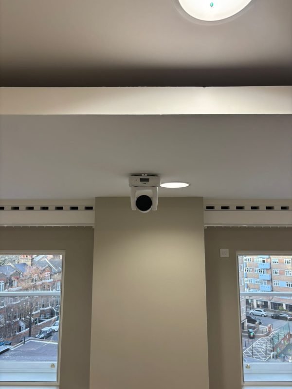 A white security camera is mounted on a ceiling near a wall, with two windows showing buildings and cars outside.