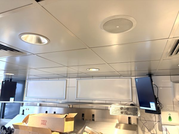 A commercial kitchen ceiling with recessed lights and speakers, a cardboard box on the counter—ready for pizza delivery—two wall-mounted monitors, and white tiled walls create the busy atmosphere of Domino’s Pizza in Hunstanton.