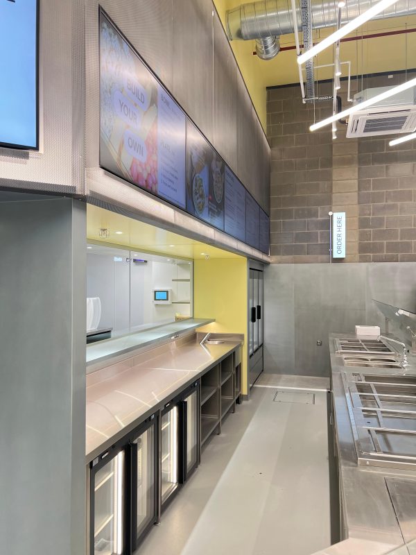 Modern restaurant counter with metal surfaces, drink coolers, and empty food stations. Digital menu boards and an Order Here sign are visible above the counter. The setting appears clean and ready for service, reflecting the contemporary style found in many London eateries such as ATIS Foods.