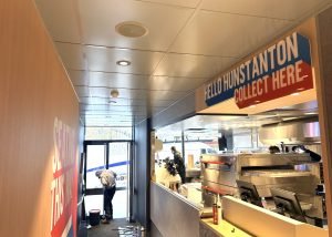 Bright, modern takeaway counter with a Hello Hunstanton Collect Here sign. A worker cleans near the door, kitchen equipment and a Domino’s Pizza delivery van outside are visible. Natural light comes in from the entrance.