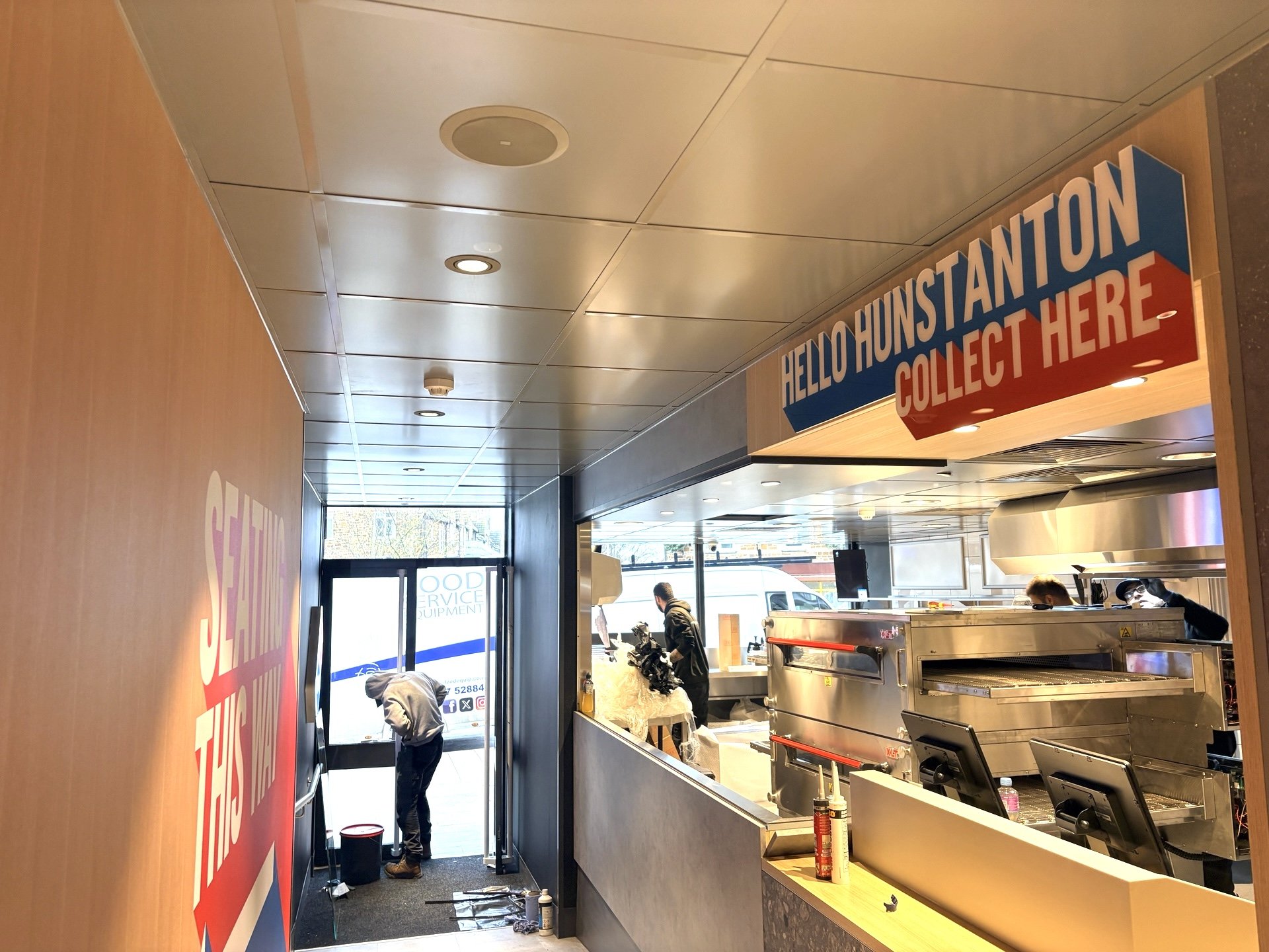 Bright, modern takeaway counter with a Hello Hunstanton Collect Here sign. A worker cleans near the door, kitchen equipment and a Domino’s Pizza delivery van outside are visible. Natural light comes in from the entrance.