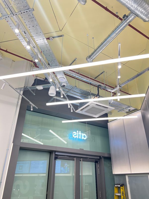 Modern office ceiling with exposed ducts, cables, and industrial light fittings, reminiscent of contemporary workspaces in London. Glass doors below reflect the lights, and a neon sign reading otis is visible through the window. A yellow ladder stands to the right, hinting at recent updates for ATIS Foods’ vibrant environment.
