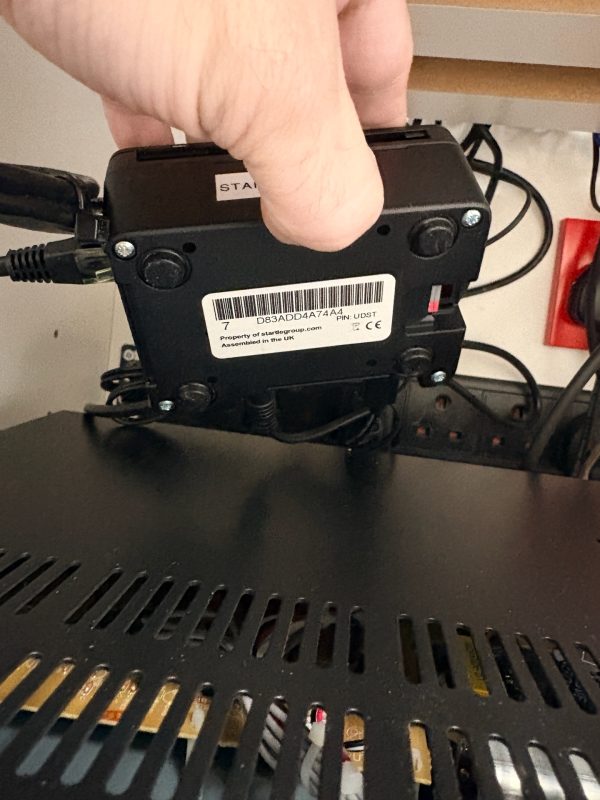 A hand is holding a black electronic device with a barcode label above a piece of equipment with vent holes, surrounded by various cables and a red electrical socket in the background—reminiscent of the organised chaos behind the scenes at Domino’s Pizza during a busy pizza delivery night in Hunstanton.