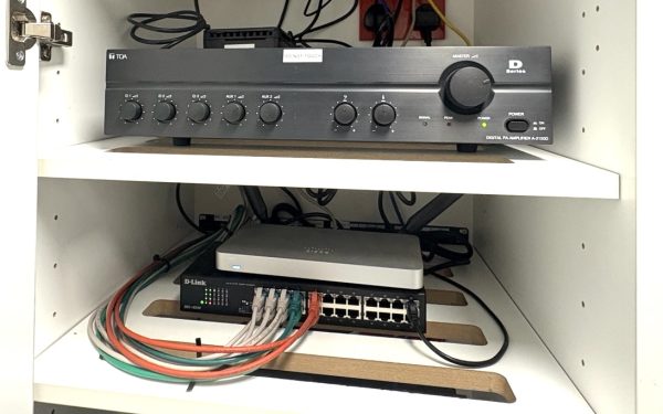 A black amplifier with dials is on the top shelf in a white cupboard, reminiscent of the clear organisation you’d expect behind the counter at Domino’s Pizza in Hunstanton. Below, a network switch with several connected Ethernet cables sits on the second shelf. The cupboard door is open.