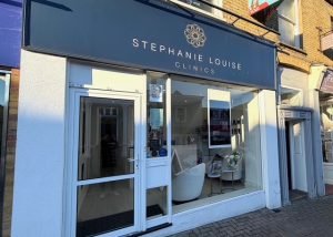 Street view of Stephanie Louise Clinics in Bishops Stortford, featuring a blue sign, glass door, and window displaying a white chair, flowers, and informational signs inside a clean, modern clinic shopfront.