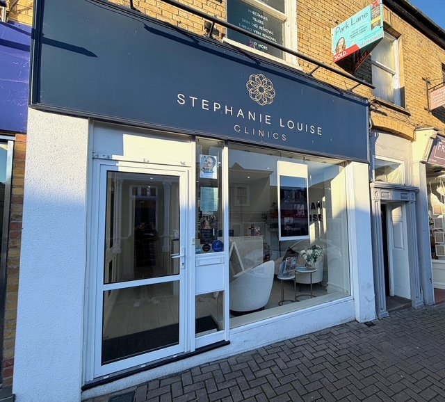 Street view of Stephanie Louise Clinics in Bishops Stortford, featuring a blue sign, glass door, and window displaying a white chair, flowers, and informational signs inside a clean, modern clinic shopfront.