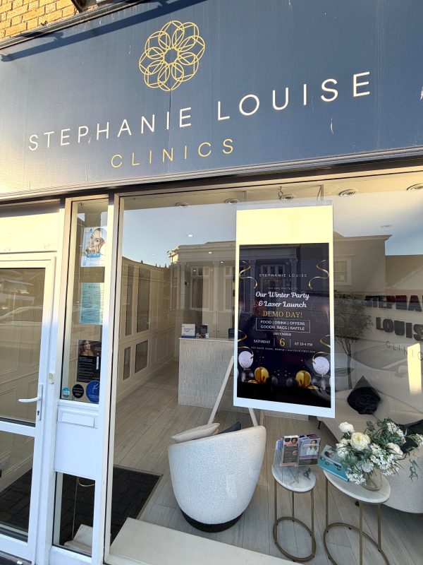 The exterior of Stephanie Louise Clinics in Bishop’s Stortford showcases a white chair, flowers, brochures, and a poster advertising a Winter Party and luxury demo day inside the window.