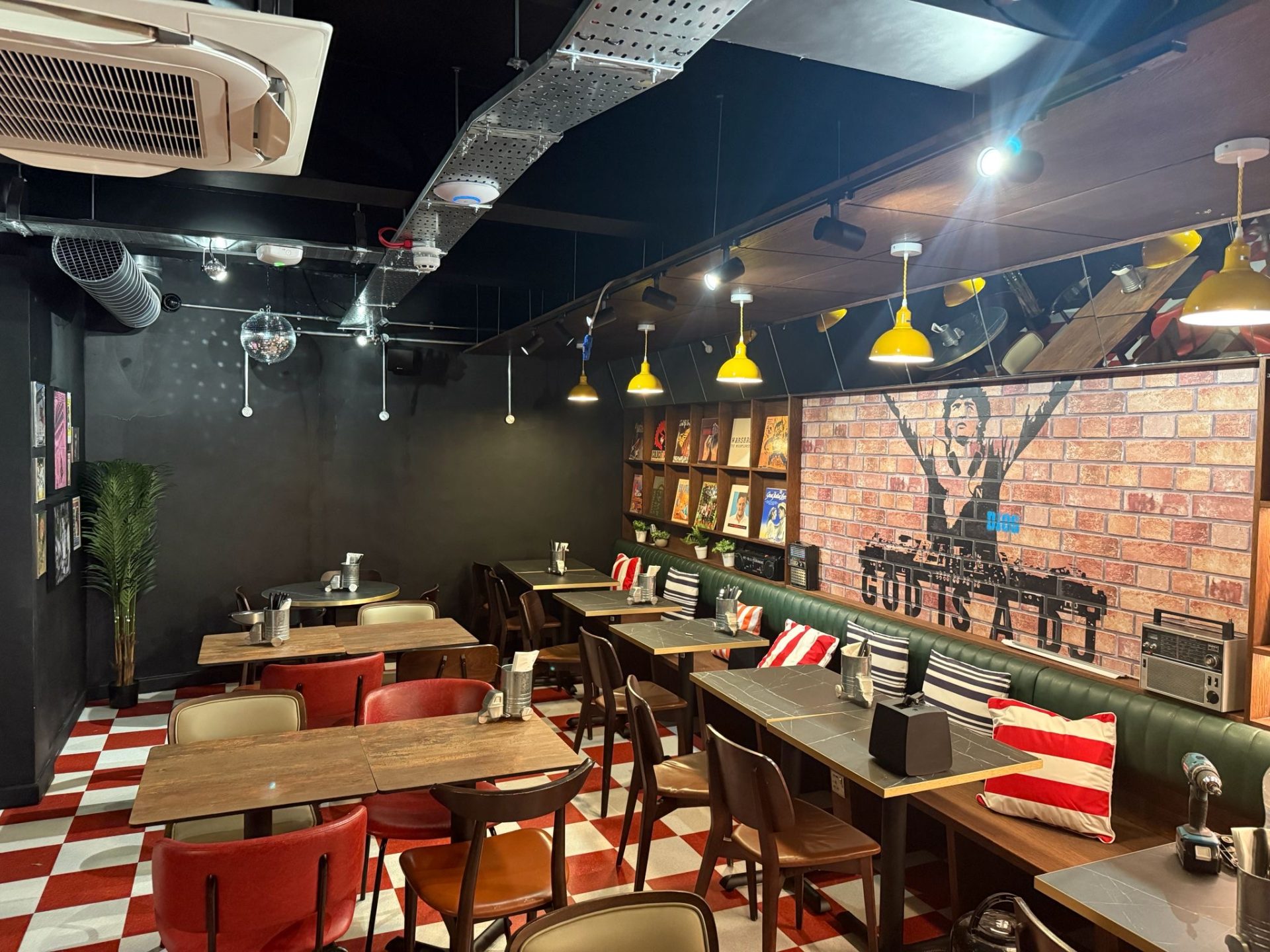 A cosy café in Bristol with wooden tables, red and brown chairs, chequered flooring, yellow hanging lights, vinyl records on shelves, and a mural of a DJ on a brick wall—a perfect spot to relax after a Pizza Pilgrims feast.