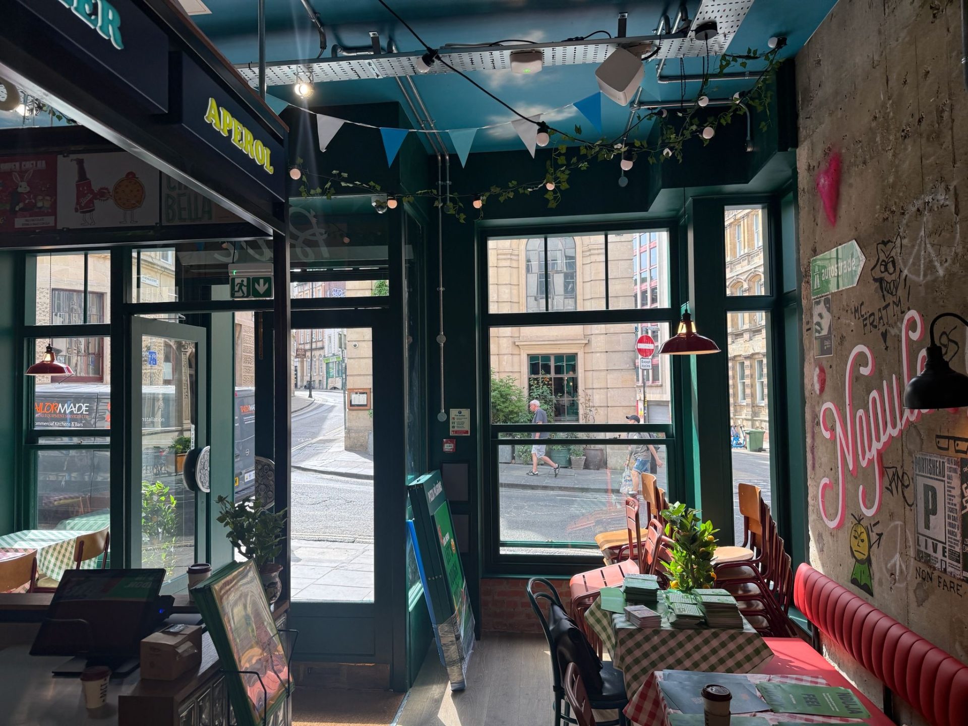 A cosy café interior with sunlight streaming through large windows, green walls, red seating, and tables set with menus welcomes guests in the heart of Bristol. Outside, a lively street and buildings are visible. Decorations and fairy lights add charm reminiscent of Pizza Pilgrims’ inviting atmosphere.