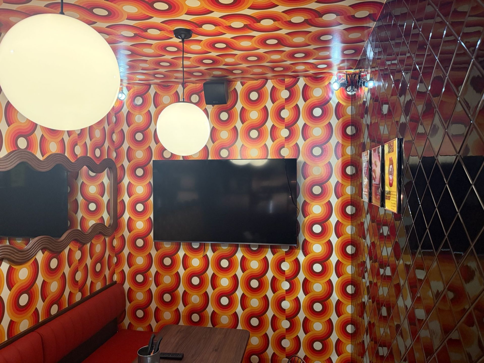 A retro-themed room with bold, circular orange and yellow patterned wallpaper, a black TV mounted on the wall, two round ceiling lights, a red bench, and a mirrored wall with a diamond pattern—this vibrant space captures the playful spirit of Pizza Pilgrims in Bristol.