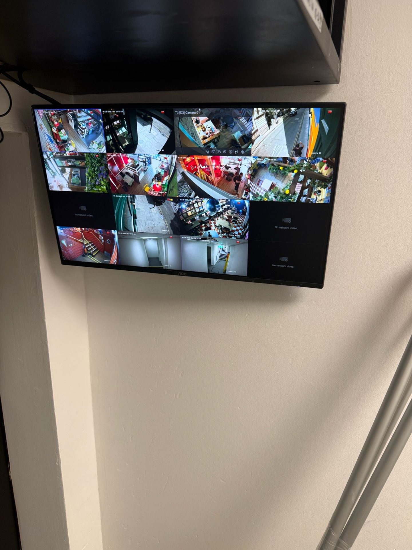 A wall-mounted monitor displays multiple CCTV feeds showing various indoor and outdoor scenes, including corridors, entrances, and a shop area. The monitor is below a black shelf and next to a light-coloured wall, providing a comprehensive view of the activities at Pizza Pilgrims in Bristol.