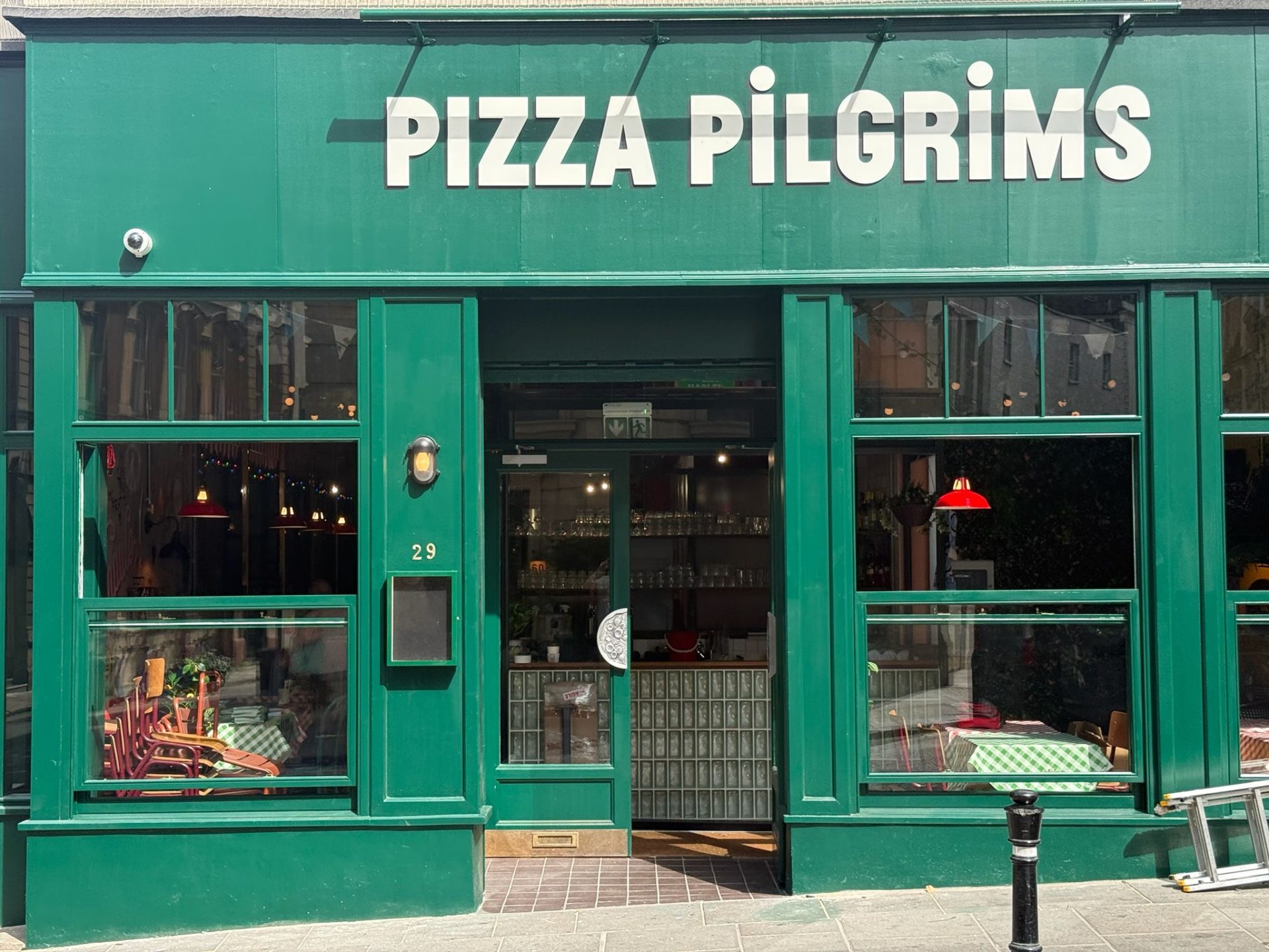 The green shopfront of Pizza Pilgrims in Bristol features large windows, a green-framed door, and the number 29 clearly visible. Stacked chairs and a table with a chequered cloth can be seen inside.