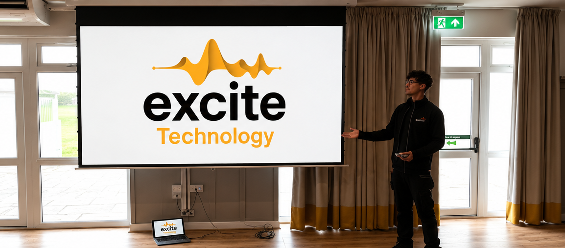 A person presents a slide with the excite Technology logo, featuring yellow sound waves, in a bright room with curtains and double doors. A laptop showing the same logo sits on the floor nearby.