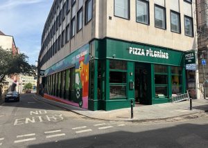 Pizza Pilgrims restaurant on a street corner in Bristol with a green exterior, large windows, colourful mural on the side wall, and a NO ENTRY sign painted on the road in front.