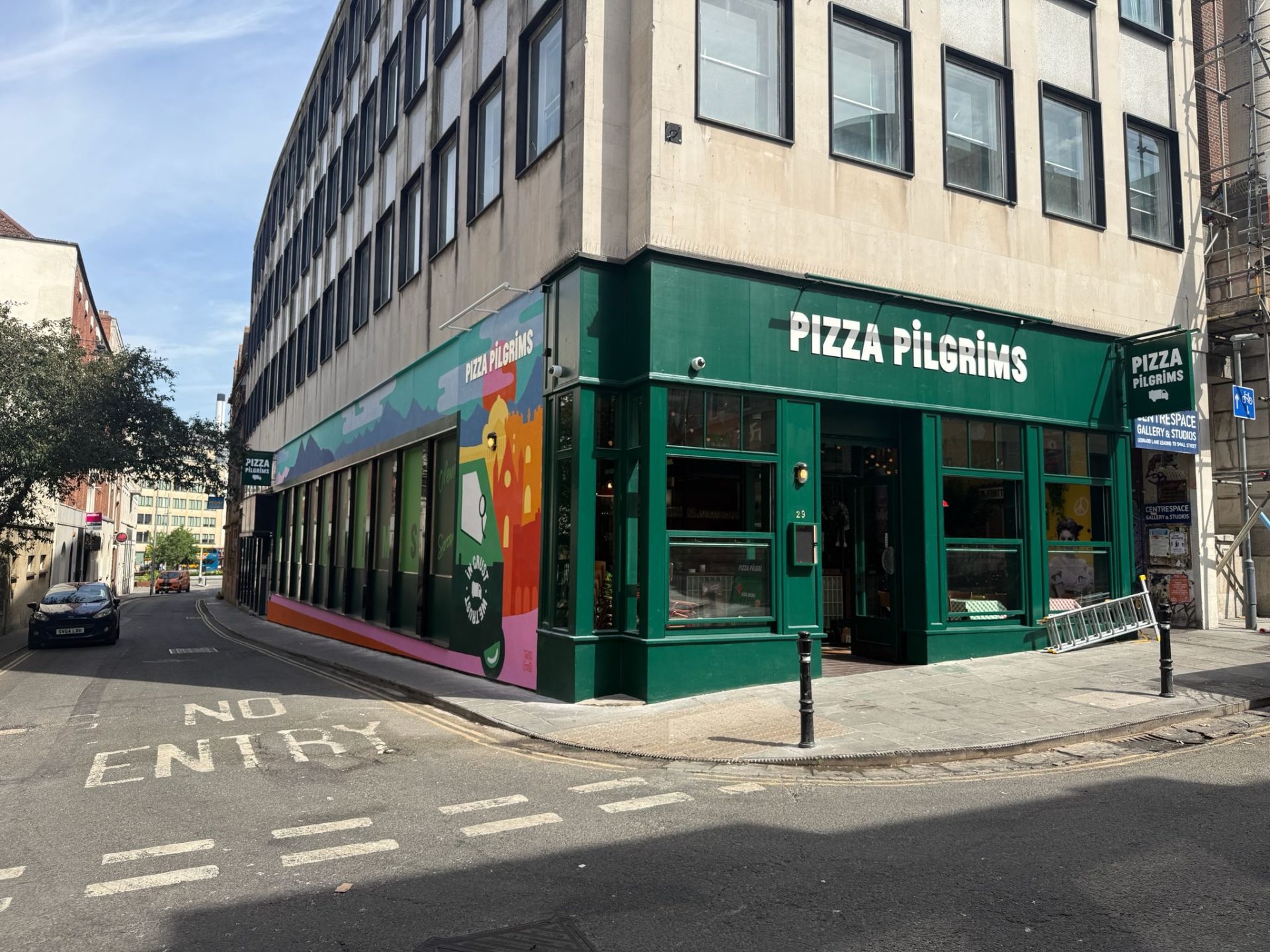 Pizza Pilgrims restaurant on a street corner in Bristol with a green exterior, large windows, colourful mural on the side wall, and a NO ENTRY sign painted on the road in front.