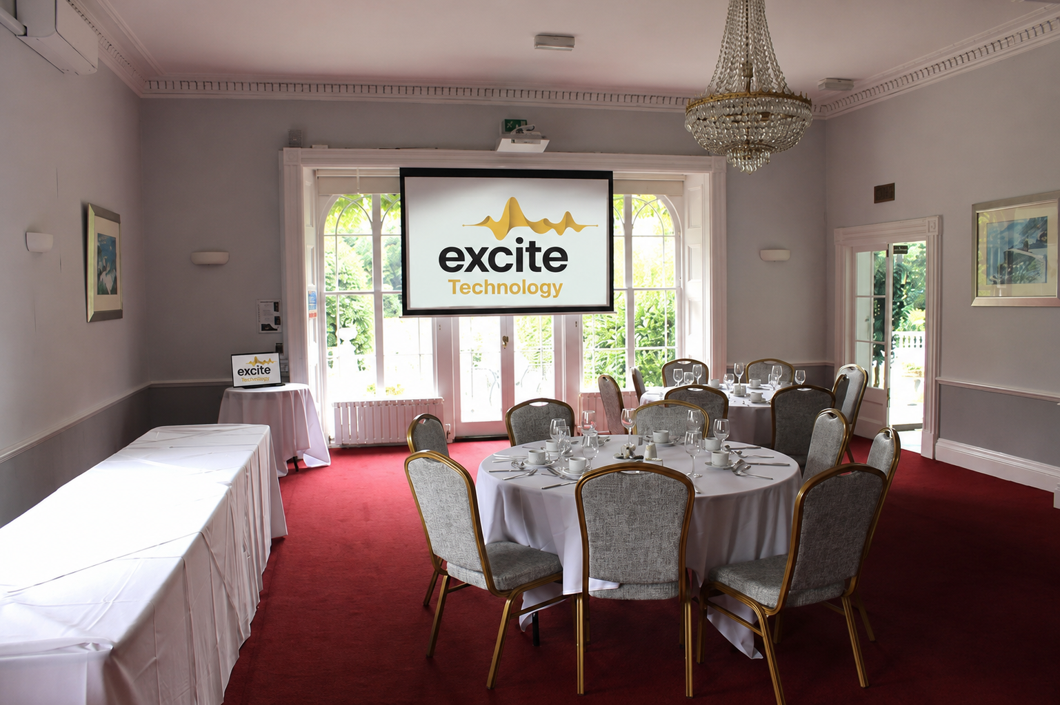 A conference room with round tables set for dining, a chandelier, a red carpet, and a projector screen displaying the excite Technology logo near large windows.