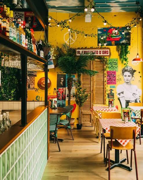 A colourful café interior in Bristol with yellow walls, framed art, leafy plants, and fairy lights. Tables have checked cloths, mismatched chairs, and vintage decor reminiscent of Pizza Pilgrims, creating a cosy, eclectic atmosphere.