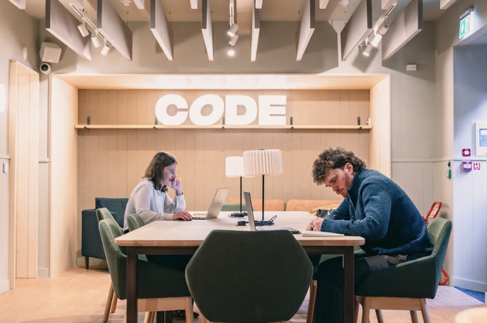 Two people work at a shared table in a modern office space in Edinburgh, with the word CODE displayed in large letters on the wall behind them, reflecting innovative Code Concepts at the heart of their collaboration.