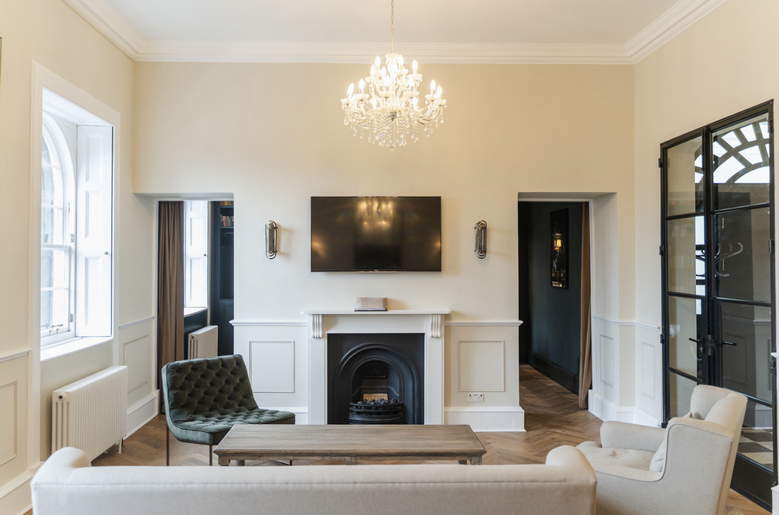 Bright living room in Edinburgh with a chandelier, wall-mounted TV above a fireplace, green and beige chairs, sofa, wooden coffee table, and large windows letting in natural light.