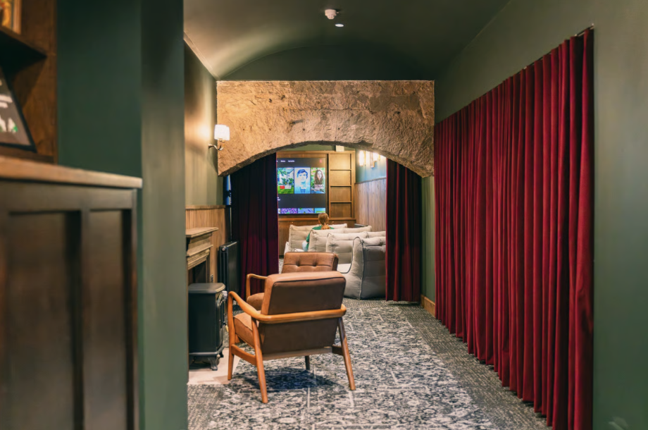 A cosy home theatre in Edinburgh features a patterned carpet, armchair, red curtains, exposed stone archway, and a sofa facing a large screen displaying film covers. Warm lighting creates an inviting atmosphere—perfect for unwinding or exploring code concepts in comfort.