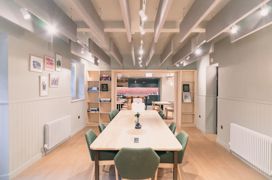A modern, brightly lit meeting room in Edinburgh with a long wooden table, green chairs, wooden shelves with books, framed art on the wall, and ceiling beams with spotlights—ideal for discussing innovative code concepts.