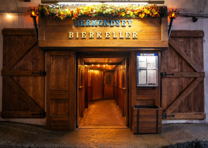 Entrance to Bermondsey Bierkeller in London with wooden doors, bright lights, autumn leaves, and a sign above. A menu is displayed on the right side by the door.