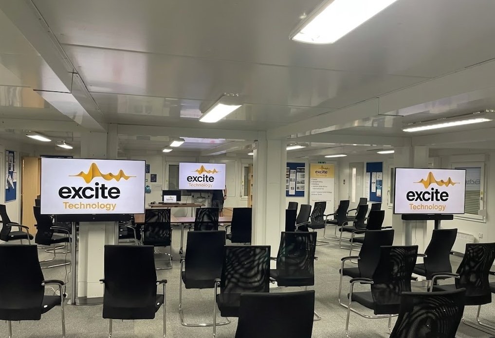 A conference room with rows of black chairs facing multiple screens displaying the excite Technology logo. The room is well-lit with white walls and ceiling.