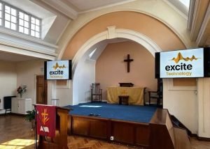 A church interior with a cross on the wall, two screens displaying Excite Technology, wooden floor, pulpit, and altar with a gold cloth. Natural light comes from large windows on the left.