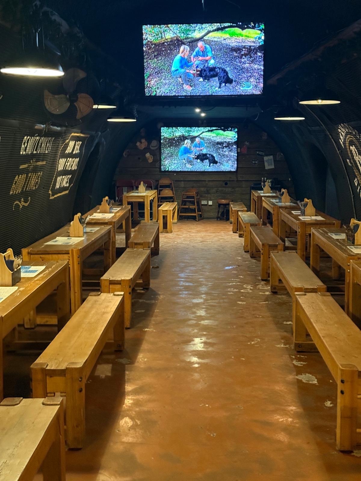 A rustic dining area with wooden tables and benches, two large screens displaying people and a dog, and dim lighting inside a tunnel-like space reminiscent of Bermondsey Bierkeller in London.
