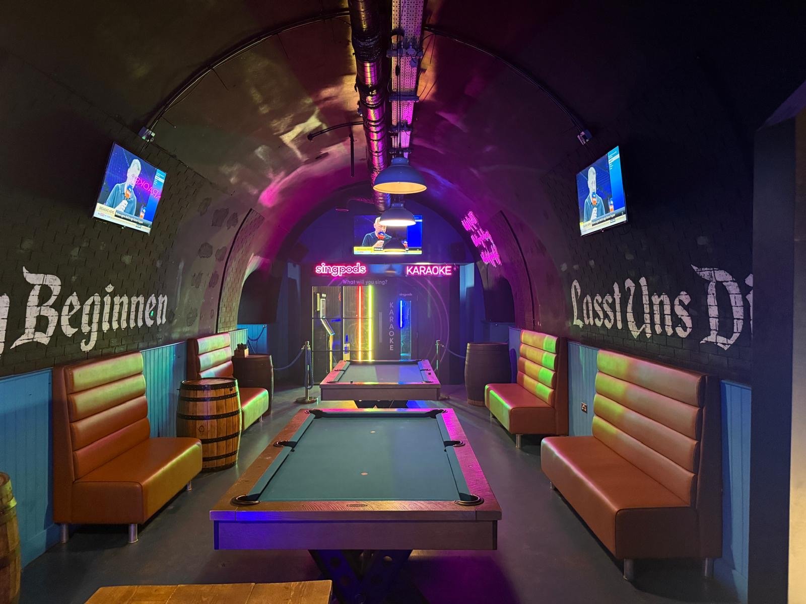 A dimly lit bar in London with arcade machines, two pool tables, leather booths, barrels as tables, neon lights, and TVs on the wall with German text on both sides captures the lively spirit of a Bermondsey Bierkeller.