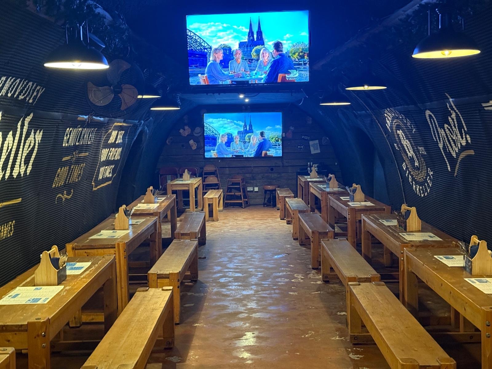A rustic indoor dining area with long wooden tables and benches, dim lighting, and two TVs on the back wall showing people dining. Menu papers and serviette holders are on the tables, capturing the cosy atmosphere reminiscent of Bermondsey Bierkeller in London.