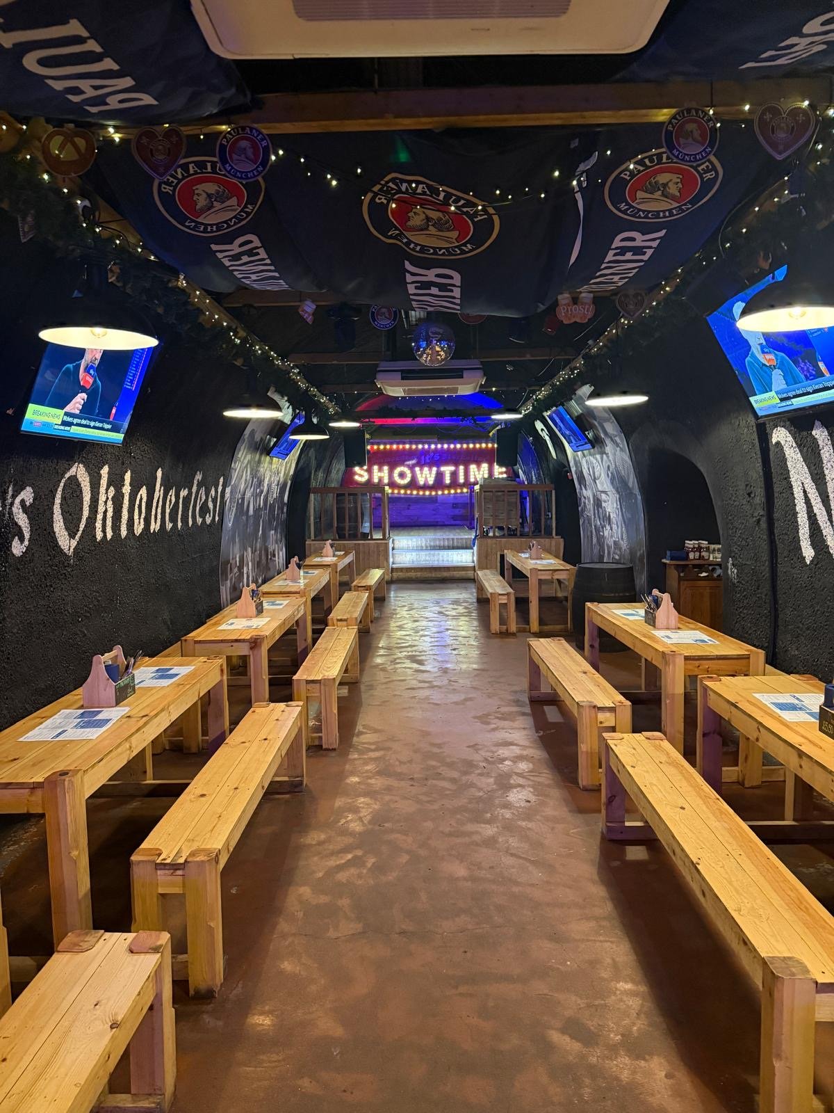 Long wooden tables and benches line both sides of a dimly lit hall with sport on TV screens, evoking the lively atmosphere of Bermondsey Bierkeller in London. A SHOWTIME sign is illuminated at the back, while decorations and flags hang from the curved ceiling.