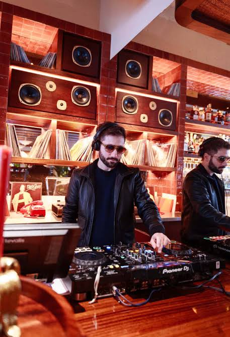 Two DJs wearing black jackets and sunglasses are mixing music at a DJ booth, surrounded by vinyl records and speakers in the stylish, retro-themed Maza Mayfair bar in London.