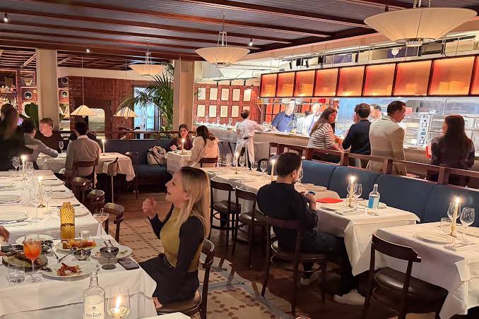 A lively restaurant scene at Maza Mayfair in London, with people dining at tables covered with white cloths, some sitting and some standing near a bar, under warm lighting and modern décor.