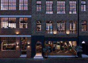 A brick building in London with large windows reveals people inside Maza Mayfair restaurant on the ground floor and offices above. Warm lights illuminate the exterior, and potted plants flank the entrance at street level.