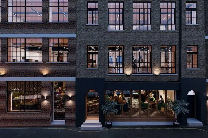 A brick building in London with large windows reveals people inside Maza Mayfair restaurant on the ground floor and offices above. Warm lights illuminate the exterior, and potted plants flank the entrance at street level.