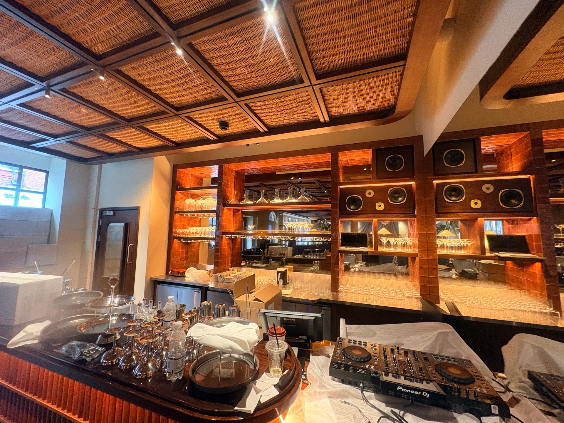 A bar at Maza Mayfair in London features shelves lined with glasses, speakers, and mirrors. The counter holds glassware, a DJ mixer, and various items, some covered with cloths. The ceiling showcases a woven pattern as sunlight streams in through the windows.