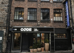 A three-storey brick building with large windows, a sign reading CODE THE LOFT, and potted plants by the entrance exudes a modern charm reminiscent of central Edinburgh. A vertical sign on the right reads CODE, highlighting the creative spirit of Code Concepts within its walls.