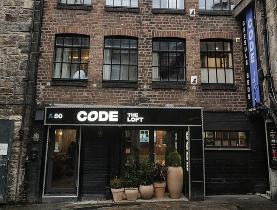 A three-storey brick building with large windows, a sign reading CODE THE LOFT, and potted plants by the entrance exudes a modern charm reminiscent of central Edinburgh. A vertical sign on the right reads CODE, highlighting the creative spirit of Code Concepts within its walls.
