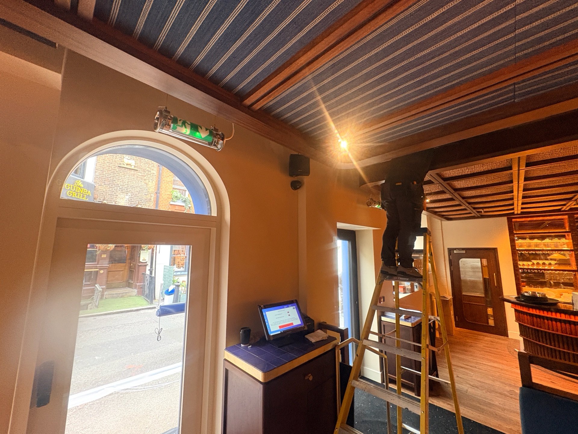 A person stands on a ladder fixing a ceiling light in Maza Mayfair, London—a restaurant with wooden beams, a striped blue ceiling, and a computer terminal near the entrance.