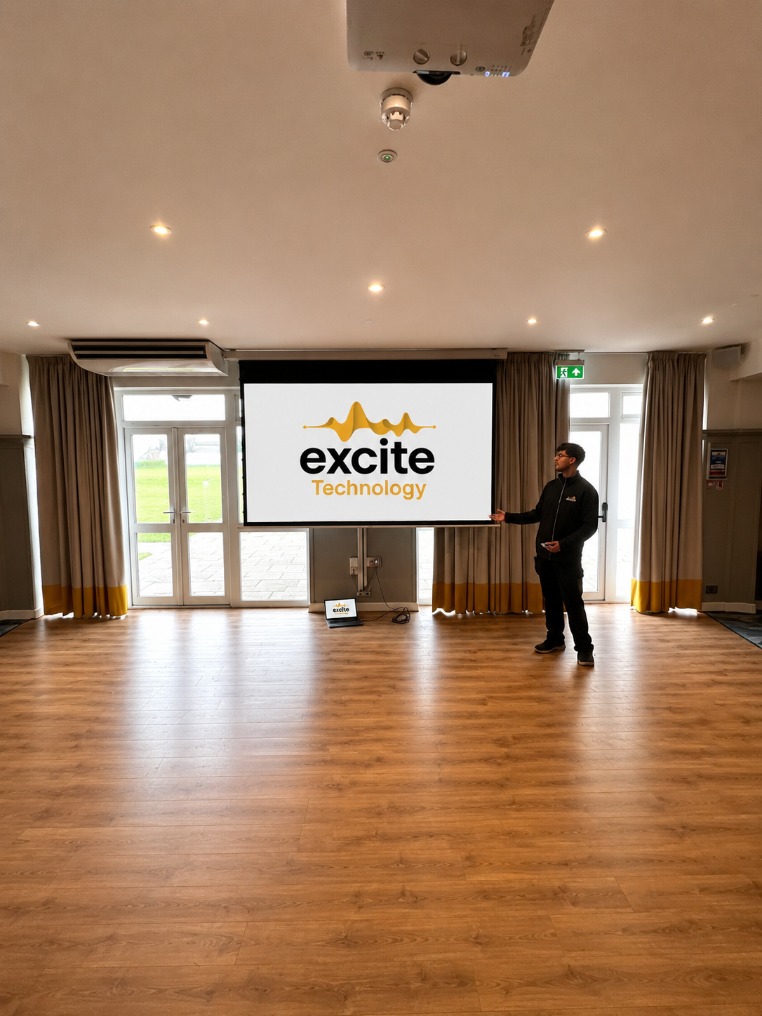 A person stands in a room pointing at a screen displaying the Excite Technology logo, with wooden floors, large windows, and curtains in the background.
