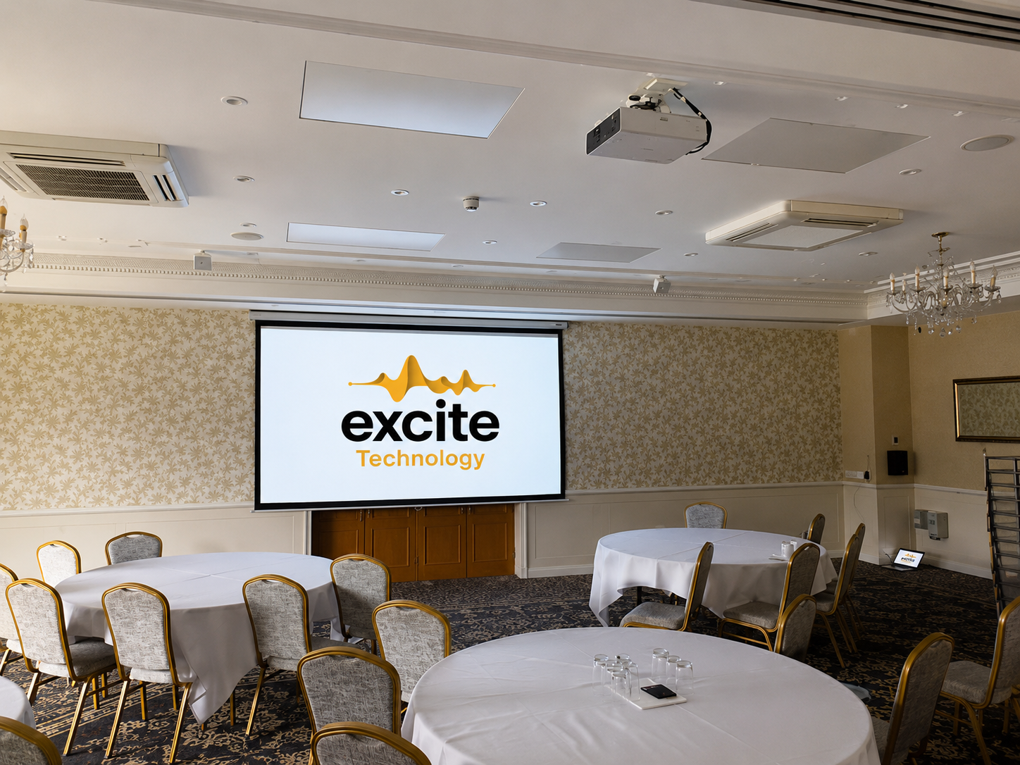Conference room with round tables, chairs, and a large screen displaying the excite Technology logo in orange and black. The room features beige wallpaper and ceiling lights.