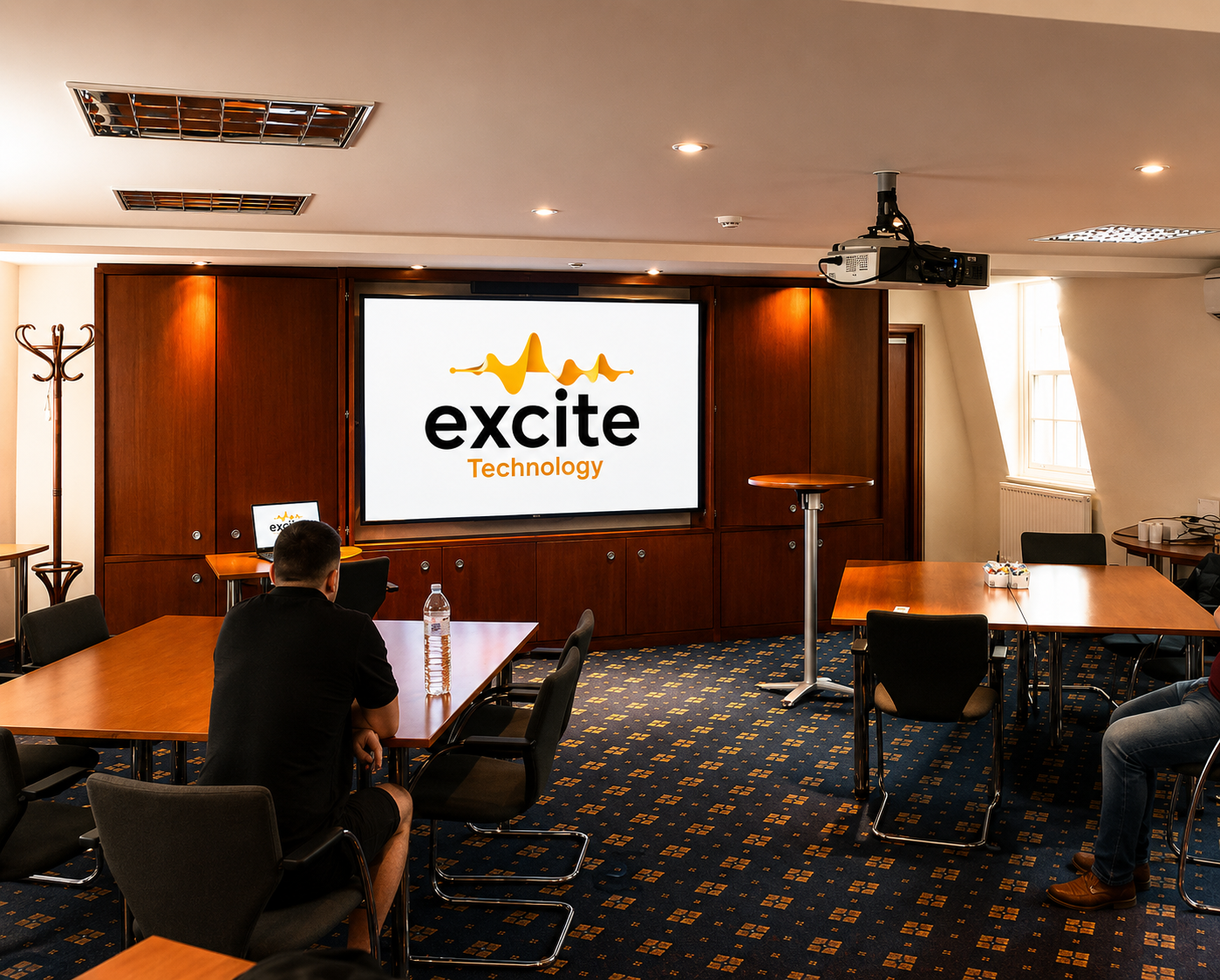 A person sits in a conference room facing a screen displaying the excite Technology logo. The room has wood panelling, tables, chairs, and a ceiling projector.