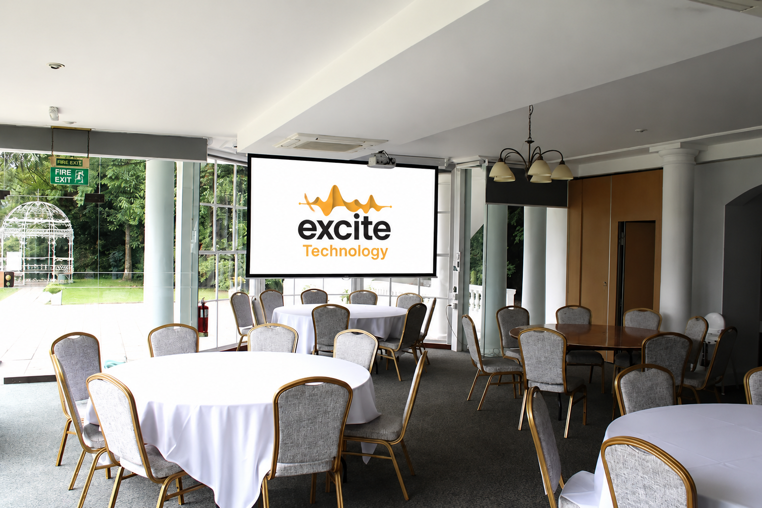 A conference room with round tables and chairs, a large screen displaying excite Technology, glass doors leading outside, and greenery visible through the windows.